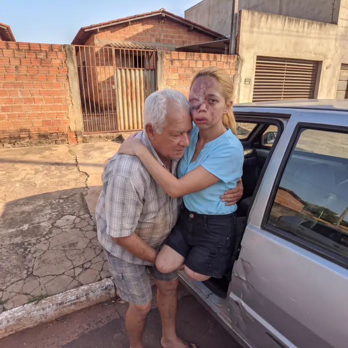 Juliana sendo carregada no colo por um homem, ao lado de um carro, na frente de casa